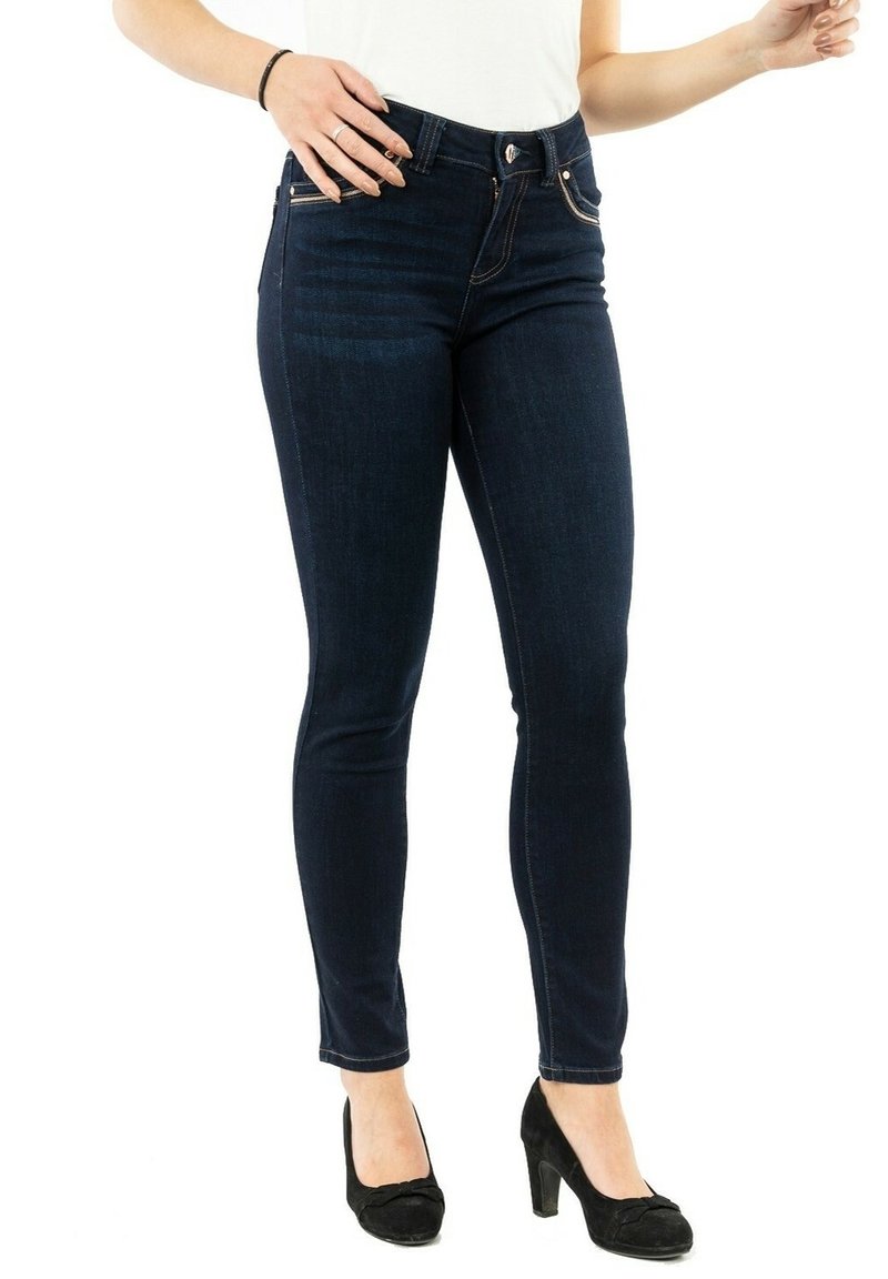 Femme portant un jean skinny bleu foncé et des chaussures à talons noirs, debout sur un fond blanc.