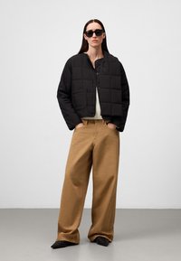 Giacca a vento trapuntata nera con un design corto, forma squadrata e due tasche frontali; abbinata a pantaloni larghi color sabbia e scarpe nere.