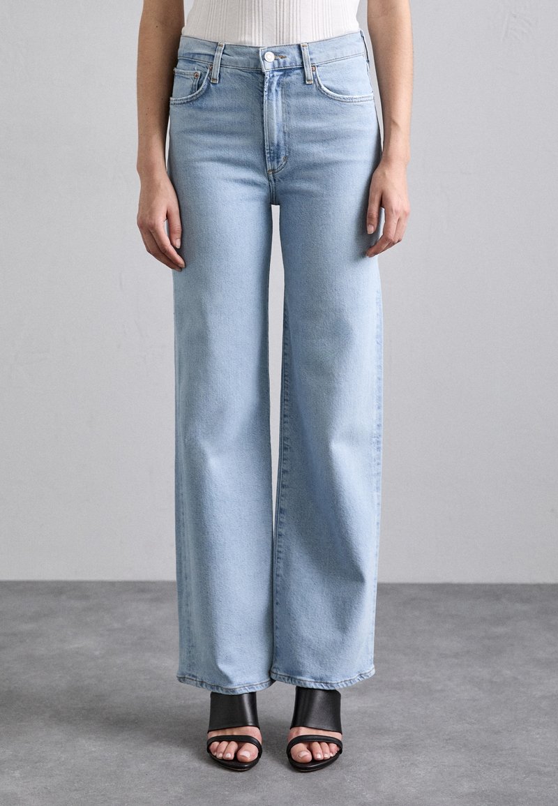 Jean bleu clair taille haute à jambes larges porté avec des sandales noires à talons ouvertes au bout et un haut blanc sur un fond gris uni au sol et au mur.