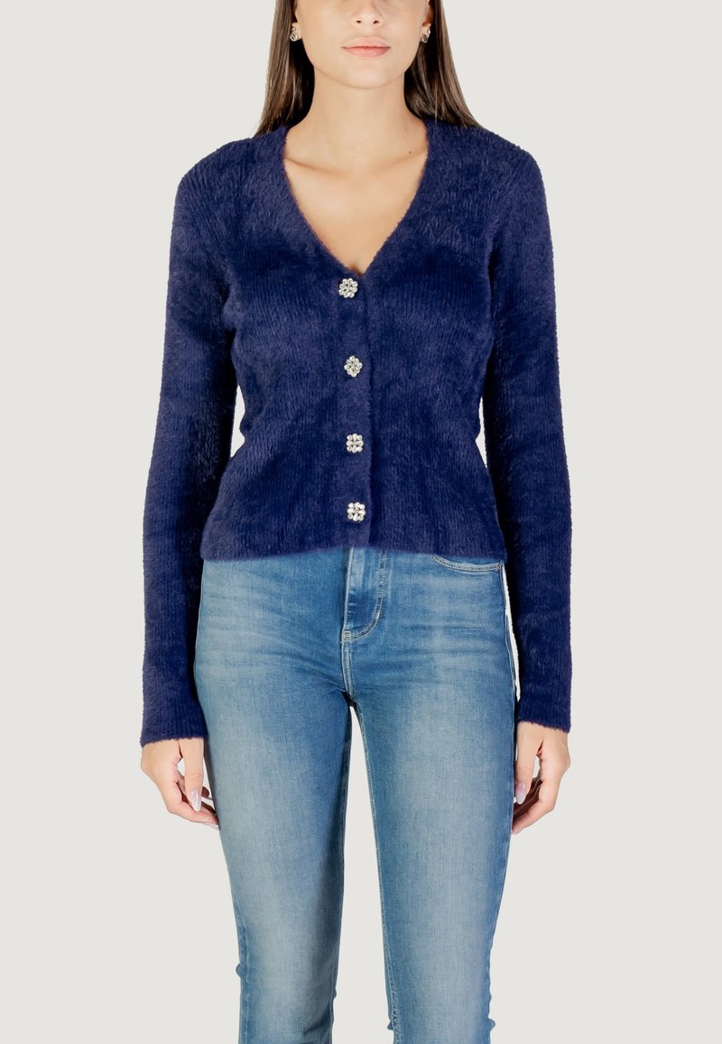 Cardigan blu navy con scollatura a V, maniche lunghe, tessuto testurizzato e cinque bottoni con motivo floreale sul davanti. Indossato con jeans azzurri chiari.