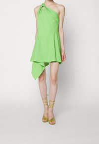 Grön enaxlad klänning i mjukt tyg, med en utsvängd kjol med en asymmetrisk fåll. Matchad med gula sandaletter med remmar.