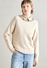 Pull en tricot beige avec une finition texturée et un col ondulé orné de embellissements transparents, associé à une chemise à col en dessous.
