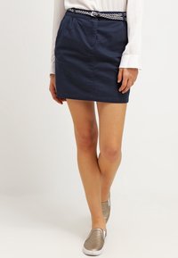 Námořnická bavlněná minisukně s poutky na opasek, která je doplněna splétaným bílým opaskem, má boční kapsy a rovný střih, spárovaná s kovovými klasickými botami na klínku.