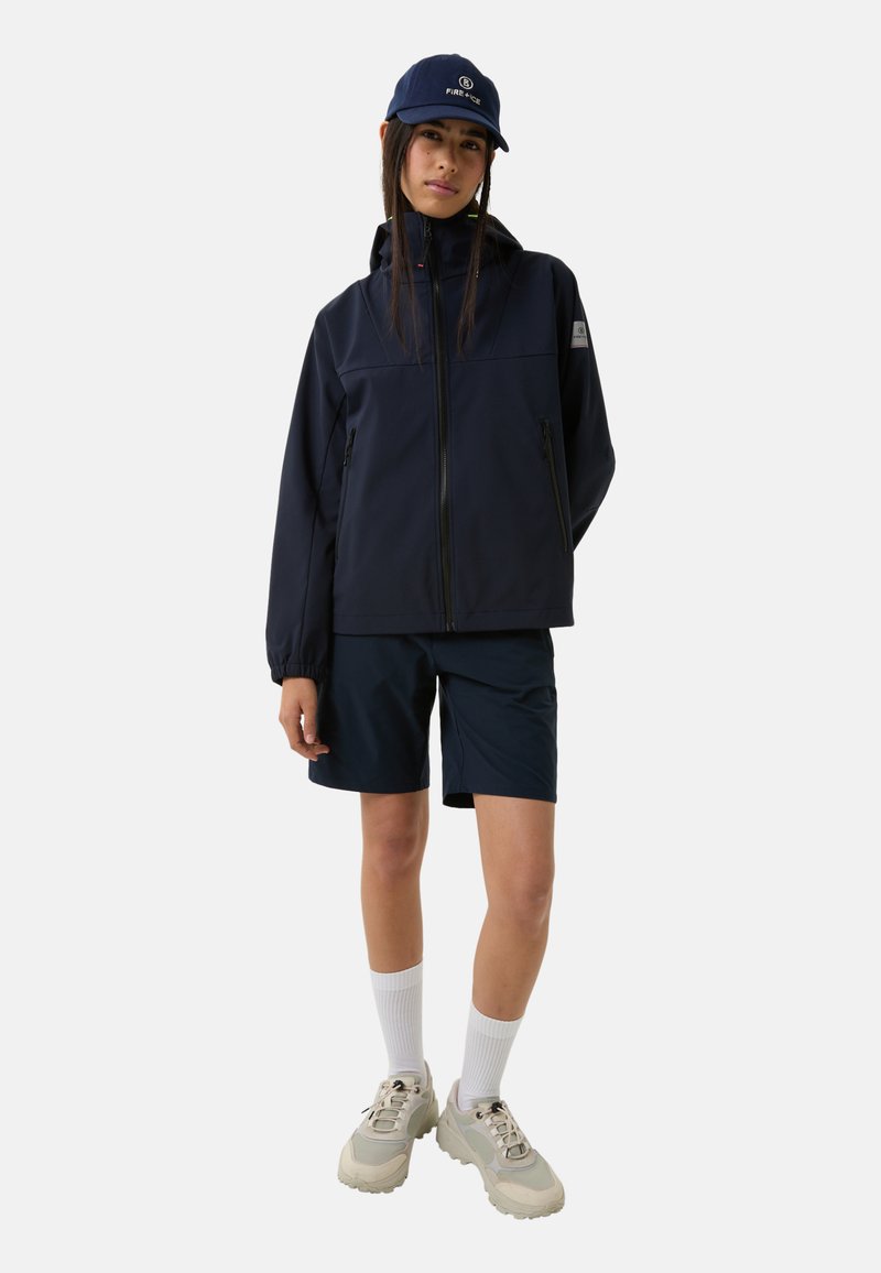 Marineblaues Jackett mit hohem Kragen und Reißverschluss, kombiniert mit marineblauen Shorts. Weiße Socken und helle klobige Sneakers vervollständigen das Outfit.
