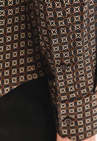 Mankiet wzorzystej koszuli z geometrycznymi brązowymi, białymi i czarnymi kształtami przypominającymi gwiazdy oraz dwoma ciemnymi guzikami na mankiecie.