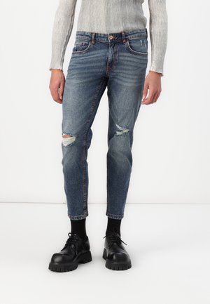 Modré džínsy z denimu so poškodenými vyvrtanými miestami, so zúženým strihom nohavíc, viditeľným šitím a zapínaním na gombík; nosené s čiernymi platformovými topánkami.