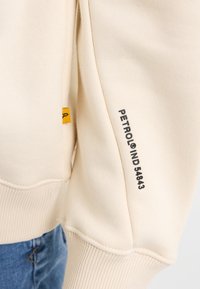Felpa in cotone beige con orlo e polsini a costine, dotata di un'etichetta gialla e testo stampato in nero sulla manica.