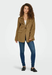 Bruine suède blazer met enkele knoopsluiting, notch revers en twee voorzakken. Gecombineerd met blauwe skinny jeans en zwarte instappers.