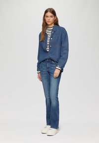 Jeanshemd, hell gewaschen, knopfgeschlossen mit zwei Brusttaschen, über einem schwarz-weiß gestreiften Langarmhemd getragen. Blaue Jeans und weiße Sneaker.