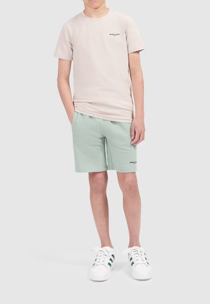 Lichtbeige t-shirt met donker logo, gecombineerd met lichtgroene shorts. Witte sneakers met groene strepen. Casual fit en zacht katoenmateriaal.