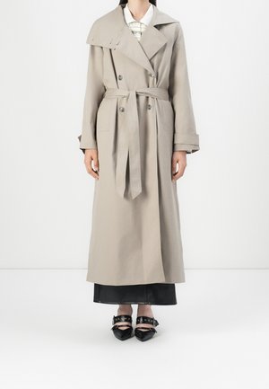 Femme portant un trench-coat beige à double boutonnage avec ceinture à la taille, jupe noire et chaussures noires pointues avec lanières et œillets métalliques.