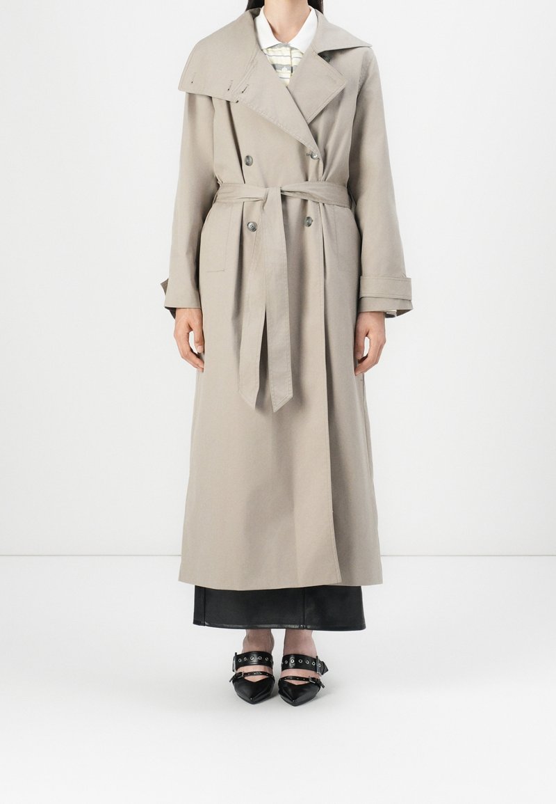 Femme portant un trench-coat beige à double boutonnage avec ceinture à la taille, jupe noire et chaussures noires pointues avec lanières et œillets métalliques.