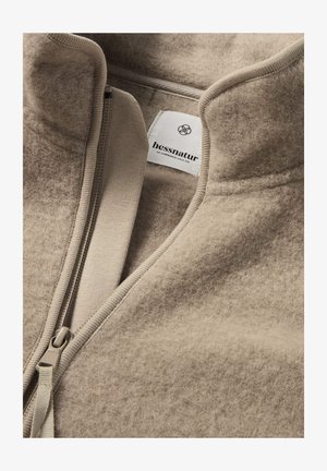 Beige Reißverschlussjacke aus weichem, angerautem Stoff. Mit Stehkragen und einem Etikett mit dem „hessnatur“ Logo.