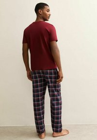 Man står barfota i en enkel burgunder t-shirt och navy, röd och vit rutiga mysbyxor mot en ljus bakgrund.