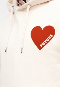 Μπεζ φούτερ με κορδόνια, με μεγάλο κόκκινο γραφικό καρδιά και τη λέξη "ΜΕΛΛΟΝ" μέσα στην καρδιά στο στήθος.