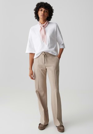 Frau mit lockigem Haar, die ein weißes, übergroßes Oberteil, beige Hosen mit weitem Bein, rosa Halstuch und braune Mokassins trägt und mit einer Hand in der Tasche steht.