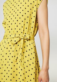 Keltainen mekko, jossa on mustia pilkkuja, lyhyet hihat ja vyötärösolmu. Kankaan pinta on sileä ja siellä on avonainen kaula-aukko.