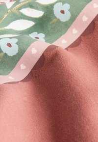 Primo piano di un tessuto rosa con bordo ondulato e motivo floreale verde, incorniciato da una striscia rosa con piccoli cuori bianchi.