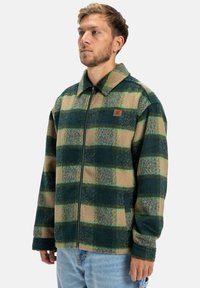 Groene en beige geruite jas met ritssluiting en grote kraag, gemaakt van gestructureerde stof. Bevat een klein leren logo-patch op de borst.