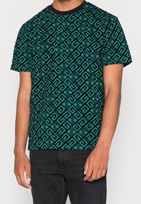 Maglietta a manica corta con scollo rotondo di colore teal, caratterizzata da un motivo geometrico nero ripetuto, realizzata in tessuto misto cotone, di peso leggero.