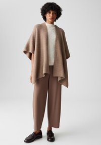 Beige ribbelgebreide poncho met wijde mouwen, gelaagd over een lichte gestructureerde sweater, gecombineerd met bruine wijde broekspijpen en donkere instappers.