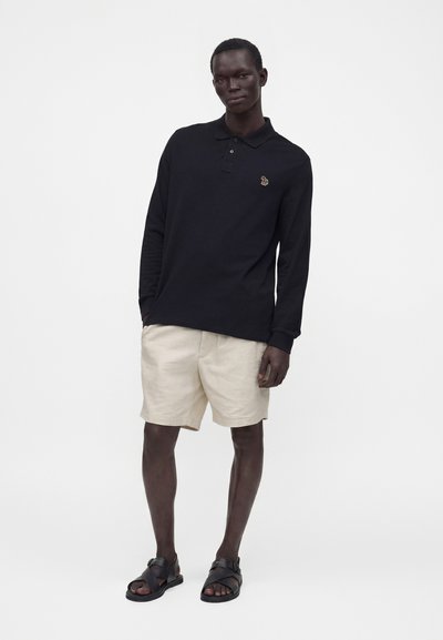 Homme debout portant un polo noir à manches longues, un short beige et des sandales noires sur un fond blanc uni.
