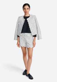 Wit en marineblauwe gestreepte jas met gouden knopen, gecombineerd met shorts met een scalloped hem. Een zwarte top en loafers maken de outfit compleet.