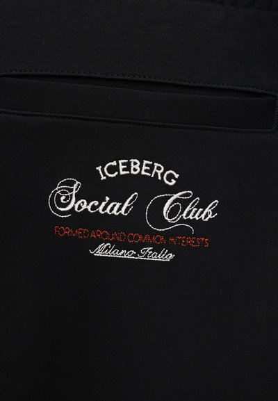 Μαύρο ύφασμα με οριζόντια σχισμή τσέπης και κεντημένο κείμενο σε λευκό και κόκκινο που αναγράφει "ICEBERG Social Club Formed Around Common Interests Milano Italia."