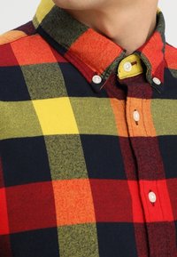 Camicia di flanella a quadri con grandi quadrati rossi, gialli, neri e verdi, colletto button-down e bottoni bianchi. Texture morbida.