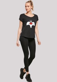 Svart T-skjorte med ansiktet til en tegneseriefigur, kombinert med svarte leggings og svarte sneakers. T-skjorten har korte ermer og en avslappet passform.