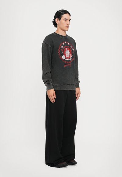 Dunkelgrauer, übergroßer Sweatshirt mit einem runden roten Grafikdruck und Text, kombiniert mit weiten schwarzen Hosen und schwarzen Schuhen.