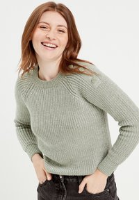 Pull en tricot vert clair avec texture côtelée, encolure ronde et boutons à l'épaule, associé à un jean noir.