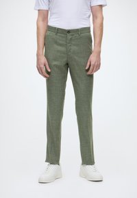 Hellgrüne Hose aus strukturiertem Stoff mit gerader Passform, ausgestattet mit einem Knopfverschluss und seitlichen Taschen, kombiniert mit weißen Turnschuhen.