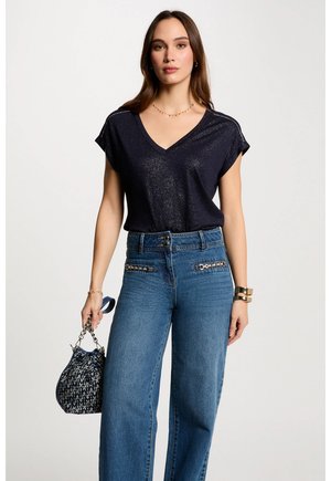 Marineblaues V-Ausschnitt-Oberteil mit glitzernder Textur und kurzen Ärmeln; kombiniert mit hochtaillierten blauen Jeans mit dekorativen Elementen. Hält eine gemusterte Handtasche.