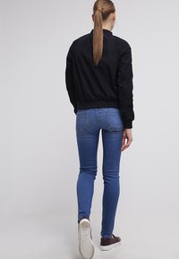 Svart bomberjacka med ribbade muddar och nederkant, i kombination med blå skinnyjeans och bruna sneakers. Modellen visas bakifrån.