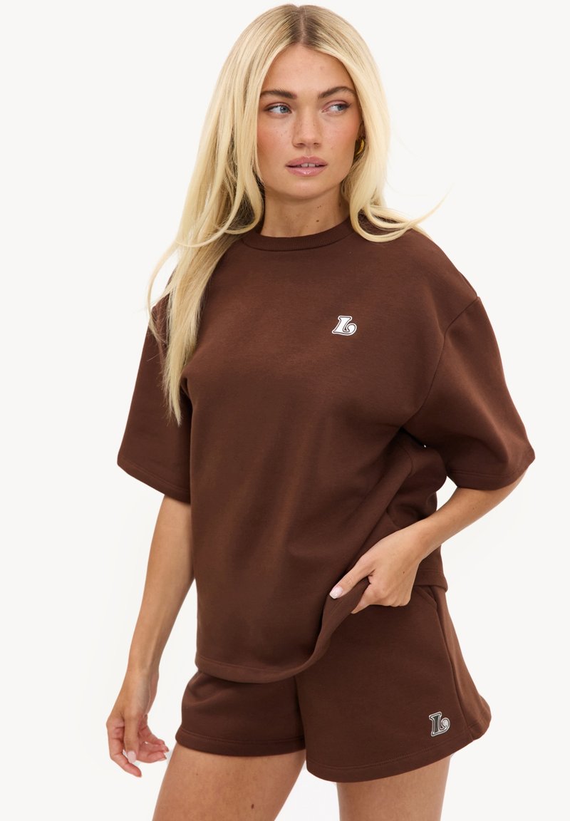 Femme aux longs cheveux blonds portant une chemise marron surdimensionnée à manches courtes et un short assorti avec de petits logos blancs.