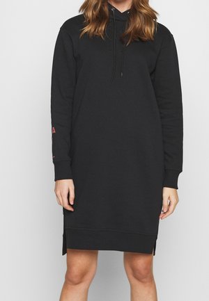 Kobieta w długiej czarnej sukience z kapturem, z rozcięciami po bokach i sznureczkami, stojąca na jednolitym białym tle.