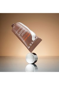 Brun kosmetiktub med vit kräm som rinner ut, balanserad på en rund metallkåpa, mot en mjuk beige bakgrund.