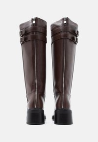 Καφέ δερμάτινες μπότες μέχρι το γόνατο με κομψό σχεδιασμό, δύο αγκράφες, φερμουάρ στο πίσω μέρος και χοντρό, υφασμένο μαύρο τακούνι.