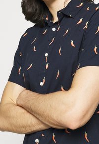 Camisa de manga corta azul marino con un patrón de chiles naranjas, botones blancos y un diseño de cuello clásico.