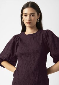 Mujer joven con pelo largo y oscuro, que lleva grandes pendientes de aro de plata y un vestido púrpura texturizado con mangas abullonadas, contra un fondo blanco.