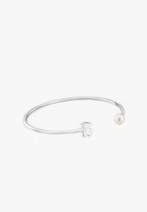 Brazalete abierto de plata con un pequeño colgante de oso de peluche en un extremo y una perla blanca en el otro extremo.