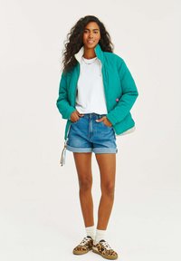 Tyrkysová izolovaná bunda so zipsom vpredu, bielym golierom a ružovým akcentom. Nosená cez biele tričko a riflové šortky s vyhrnutými manžetami. Topánky s leopardím vzorom.