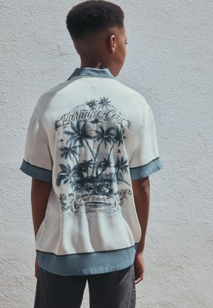 Jong persoon staat tegen een lichte muur, draagt een wit-blauw shirt met een zwart palmboomdesign en "Paradise Cove" tekst op de achterkant.