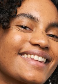 Nærbilde av et smilende ansikt med middels brun hud, krøllete hår og naturlig sminke. Kjennetegn inkluderer klar hud, veldefinerte øyenbryn og lys rosa lepper.