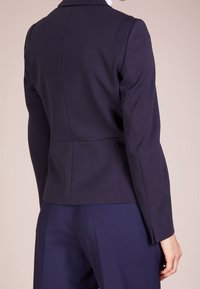 Blazer sartoriale blu navy, design strutturato, texture liscia, vestibilità slim, spacco singolo sul retro e maniche lunghe. Abbinato a pantaloni blu navy.