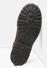 Suela de bota de goma marrón con surcos profundos para tracción, que presenta un logo circular y patrones de cruz distintivos, destacando la durabilidad y el agarre.