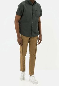 Camicia a maniche corte verde scuro con bottoni e tasca sul petto, abbinata a pantaloni cargo marrone chiaro con tasche laterali e sneakers bianche.