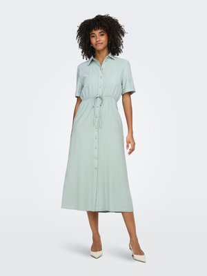Vrouw met krullend haar die een lichtgroene midi-jurk met knopen en koord in de taille draagt en witte puntige hakken, staand tegen een effen achtergrond.