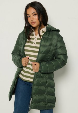 Femme portant une longue doudoune verte par-dessus un polo rayé vert et beige et un jean bleu, tenant la fermeture éclair de la veste.